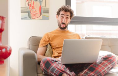 Genç sakallı adam tiksinmiş ve rahatsız olmuş hissediyor, dilini dışarı çıkarıyor, iğrenç ve iğrenç bir şeyden hoşlanmıyor ve dizüstü bilgisayarla oturuyor.