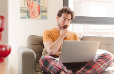 Genç sakallı bir adam ağzı ve gözleri açık ve eli çenesinde, hoş olmayan bir şok geçiriyor, ne ya da vay canına diyor ve dizüstü bilgisayarla oturuyor.