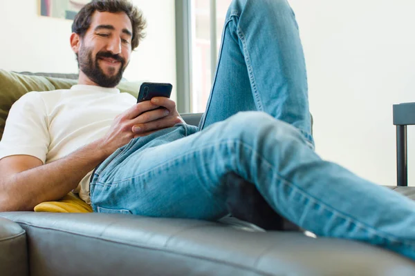 Genç sakallı adam akıllı bir telefonla kanepede dinleniyor.
