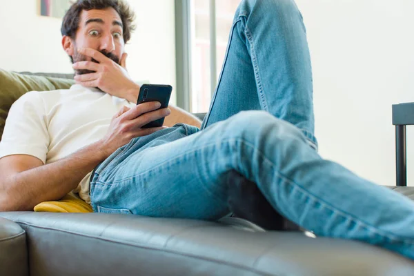 Genç sakallı adam akıllı bir telefonla kanepede dinleniyor.