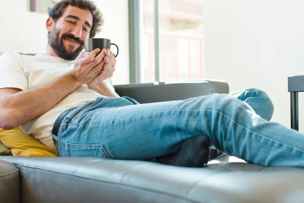 Genç sakallı adam elinde kahve fincanıyla kanepede dinleniyor.