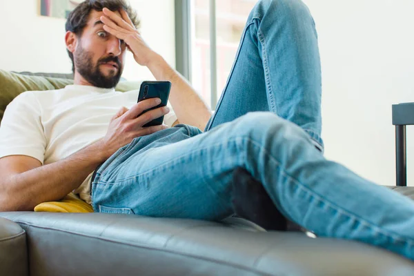 Genç sakallı adam akıllı bir telefonla kanepede dinleniyor.