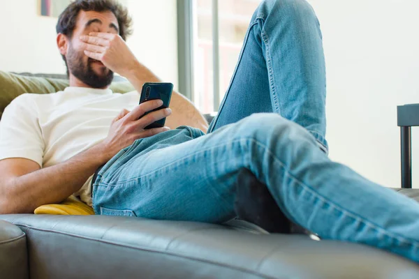 Genç sakallı adam akıllı bir telefonla kanepede dinleniyor.