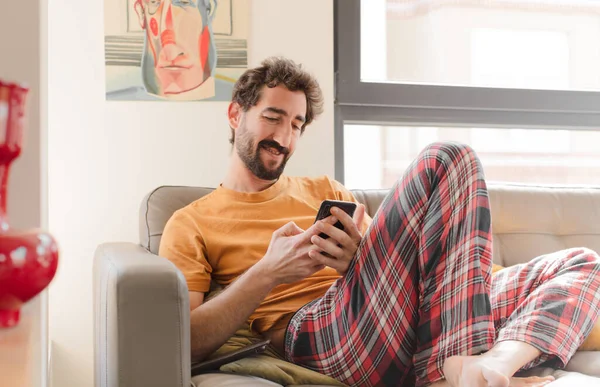 Genç sakallı adam akıllı bir telefonla kanepede oturuyor.