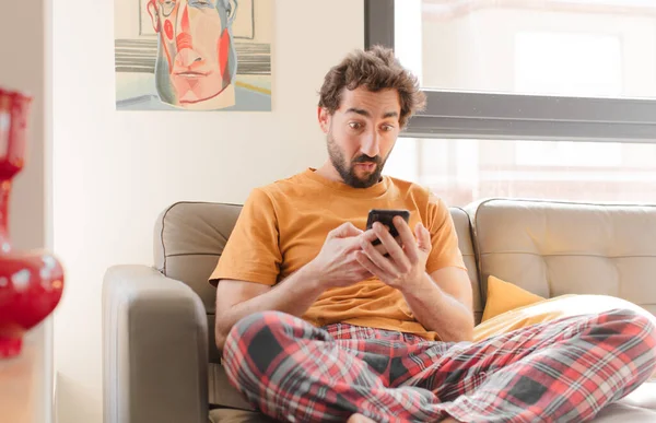 Genç sakallı adam akıllı bir telefonla kanepede oturuyor.