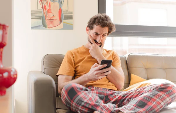 Genç sakallı adam akıllı bir telefonla kanepede oturuyor.