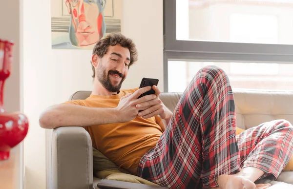 Genç sakallı adam akıllı bir telefonla kanepede oturuyor.