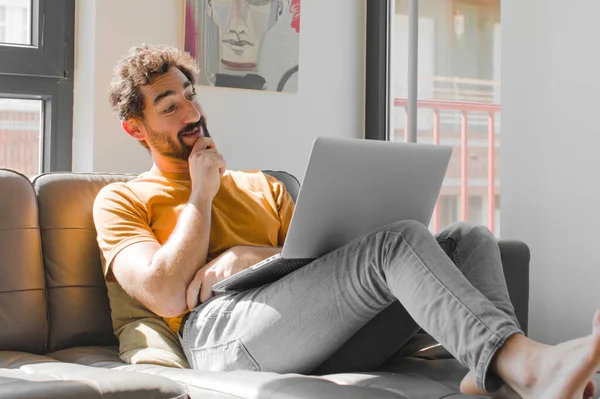 Genç sakallı adam mutlu bir şekilde gülümsüyor ve hayal kuruyor ya da şüphe ediyor, dizüstü bilgisayarla kanepe konseptine bakıyor.