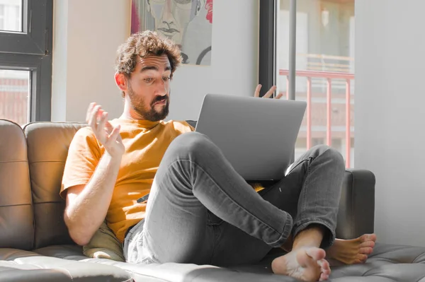 Genç sakallı adam şaşkın ve şaşkın hissediyor, kuşkucu, farklı seçenekleri tartıyor ya da değerlendiriyor kanepe dizüstü bilgisayarındaki dizüstü bilgisayarla komik bir ifadeyle.