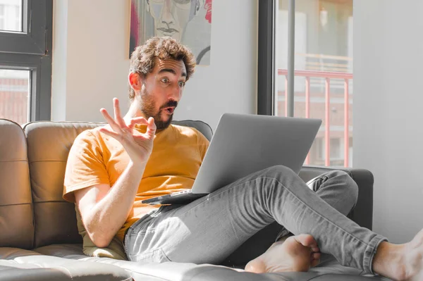 Genç sakallı adam mutlu, rahat ve tatmin olmuş hissediyor. İyi bir jest yapıyor. Dizüstü bilgisayarla kanepe konseptinde gülümsüyor.