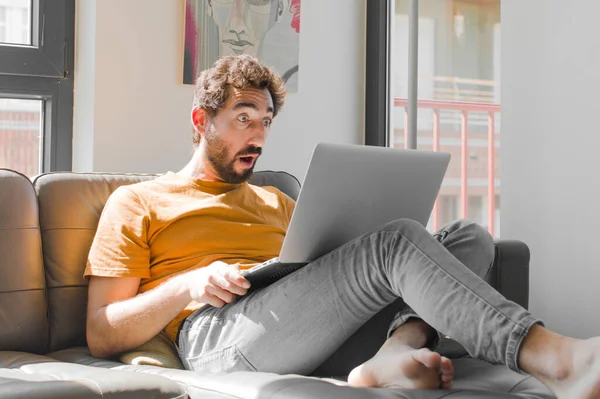 Genç sakallı adam şok olmuş ya da şaşırmış görünüyor, ağzı açık bir şekilde dizüstü bilgisayarla kanepe konseptinde 