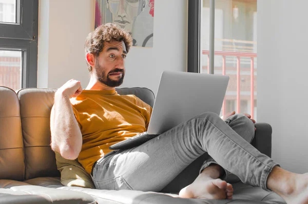 Genç sakallı bir adam stresli, endişeli, yorgun ve hayal kırıklığına uğramış, gömlek boyunlu, kanepe dizüstü bilgisayarındaki bir dizüstü bilgisayardan rahatsız olmuş gibi görünüyor.