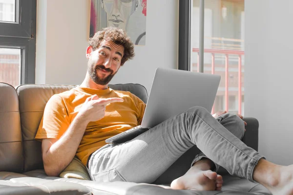 Genç sakallı adam neşeyle gülümsüyor, mutlu hissediyor ve yan ve yukarı doğru işaret ediyor, kopya uzayda dizüstü bilgisayarla kanepe dizüstü bilgisayar konseptinde nesne gösteriyor.