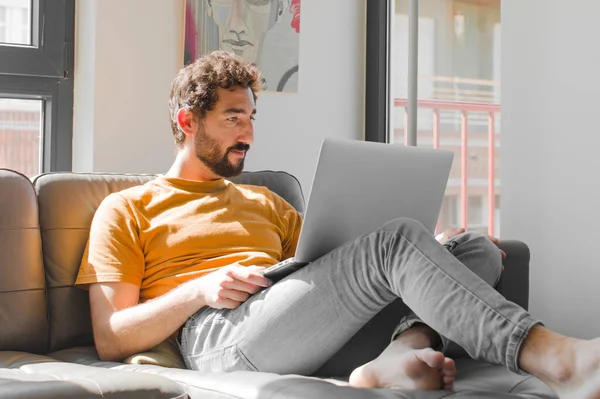 young bearded man on profile view looking to copy space ahead, thinking, imagining or daydreaming with a laptop on a couch laptop concept