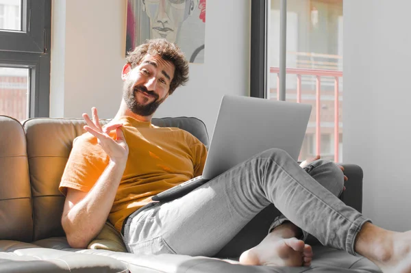 Genç sakallı adam kibirli, başarılı, pozitif ve gururlu görünüyor. Dizüstü bilgisayarla kanepe konseptini işaret ediyor.