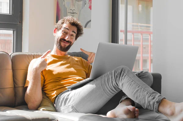 Genç sakallı adam kendi geniş gülümsemesine güvenerek gülümsüyor. Kanepe dizüstü bilgisayarındaki dizüstü bilgisayarla pozitif, rahat, tatmin olmuş bir tavır sergiliyor.