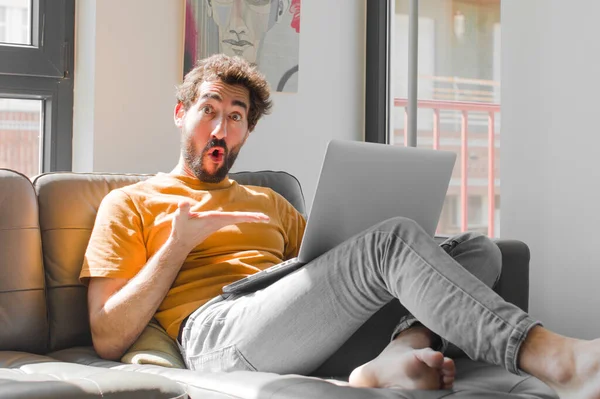 Genç sakallı adam şaşırmış ve şok olmuş görünüyor, çenesi düşmüş, eli açık bir objeyi elinde tutuyor kanepe dizüstü bilgisayarında bir dizüstü bilgisayarla