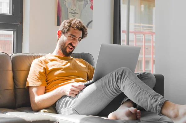 young bearded man feeling puzzled and confused, with a dumb, stunned expression looking at something unexpected with a laptop on a couch laptop concept