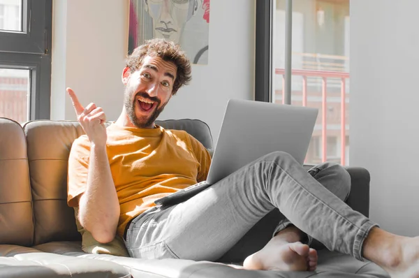 Genç sakallı adam bir fikri fark ettikten sonra mutlu ve heyecanlı bir dahi gibi hissediyor, neşeli bir şekilde parmağını kaldırıyor, eureka! Dizüstü bilgisayarlı kanepe konsepti.