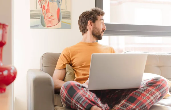 young bearded man on profile view looking to copy space ahead, thinking, imagining or daydreaming and sitting with a laptop