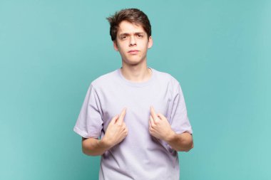 Genç bir çocuk şaşkın ve şaşkın bir bakışla kendini işaret ediyor. Seçildiğine şaşırmış ve şaşırmış.