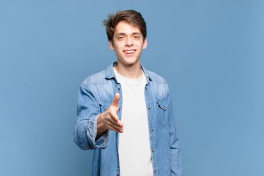 Genç çocuk gülümsüyor, mutlu görünüyor, kendinden emin ve arkadaş canlısı, bir anlaşmaya varmak için el sıkışma teklif ediyor, işbirliği yapıyor.