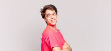 Genç çocuk sevinçle gülümsüyor, mutlu, tatmin olmuş ve rahatlamış, çapraz kollarıyla yan tarafa bakıyor.