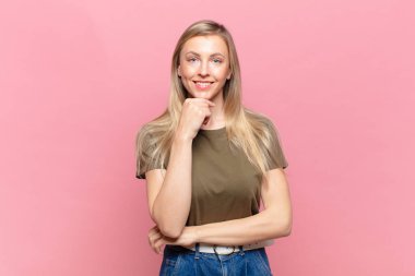 Genç sarışın güzel kadın mutlu görünüyor ve gülümsüyor. Elleri çenesinde, merak ediyor ya da soru soruyor. Seçenekleri karşılaştırıyor.