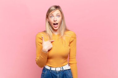 Genç, sarışın, güzel bir kadın ağzı açık, şaşkın ve şaşkın bir şekilde kendini işaret ediyor.