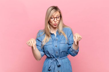 Genç, sarışın, ağzı açık, iki eliyle aşağıya doğru bakan, şaşkın, şaşkın ve şaşırmış görünen güzel bir kadın.