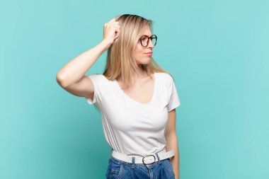 Genç sarışın güzel kadın şaşkın ve şaşkın hissediyor, başını kaşıyor ve yan tarafa bakıyor.