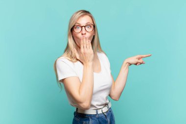 Genç sarışın güzel kadın mutlu, şok olmuş ve şaşırmış hissediyor. Ağzını eliyle kapatıyor ve yanal fotokopi alanını işaret ediyor.
