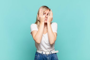 Sarışın, güzel bir kadın elleriyle yüzünü kapatıyor. Şaşırmış bir ifadeyle parmaklarının arasından bakıyor ve yan tarafa bakıyor.