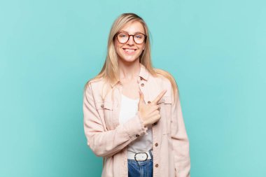 Genç sarışın güzel kadın neşeyle gülümsüyor, mutlu hissediyor ve yan ve yukarı doğru işaret ediyor, fotokopi uzayındaki nesneleri gösteriyor.