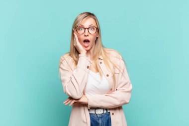 Genç sarışın, güzel bir kadın şok ve inançsızlıkla ağzı açık, elleri yanağında ve kolları çapraz, sersemlemiş ve şaşırmış hissediyor.