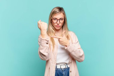 Genç sarışın güzel kadın sabırsız ve kızgın görünüyor, saati gösteriyor, dakiklik istiyor, zamanında gelmek istiyor.