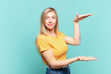 Genç, sarışın, güzel bir kadın iki eli yanda bir objeyi tutuyor fotokopi alanında, bir objeyi gösteriyor, sunuyor ya da reklamını yapıyor.