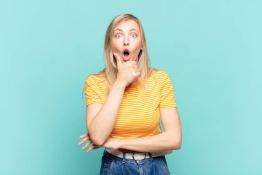 Genç, sarışın, ağzı ve gözleri açık ve eli çenesinde olan güzel bir kadın. Hoş olmayan bir şok geçiriyor.