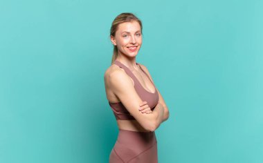 Genç sarışın kadın kameraya çapraz kollarıyla gülümsüyor ve mutlu, kendinden emin, tatmin olmuş bir yüz ifadesi var. spor konsepti