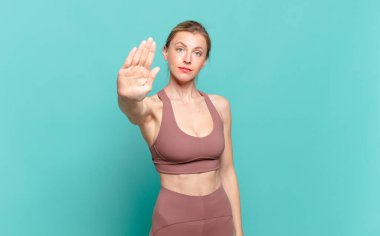 Genç sarışın kadın ciddi, haşin, hoşnutsuz ve kızgın görünüyor. Avuç içi açık bir şekilde durup jest yapıyor. spor konsepti