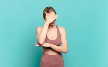 Genç sarışın kadın stresli, utanmış ya da üzgün görünüyor, baş ağrısı var, yüzünü eliyle kapatıyor. spor konsepti