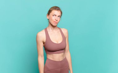 Genç sarışın kadın şaşkın ve şaşkın görünüyor, merak ediyor ya da bir sorunu çözmeye çalışıyor ya da düşünüyor. spor konsepti