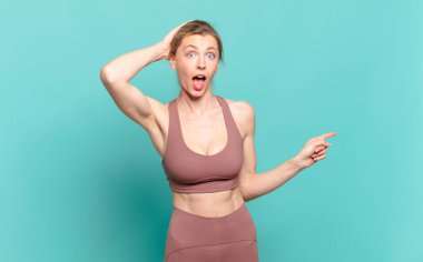 Genç sarışın kadın gülüyor, mutlu, pozitif ve şaşırmış görünüyor, yanal fotokopi alanını işaret eden harika bir fikrin farkına varıyor. spor konsepti