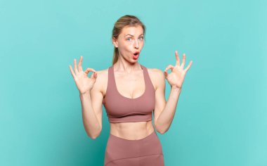 Genç sarışın kadın şok olmuş, şaşırmış ve şaşırmış hissediyor. Onayladığını gösteriyor. İki eliyle de onay işareti yapıyor. spor konsepti