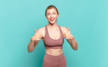 young blond woman smiling broadly looking happy, positive, confident and successful, with both thumbs up. sport concept clipart