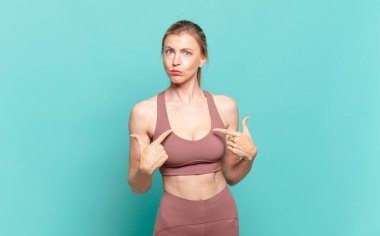 Genç sarışın kadın şaşkın ve şaşkın bir bakışla kendini işaret ediyor. Seçildiğine şaşırmış ve şaşırmış. spor konsepti