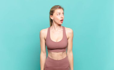 Genç sarışın kadın şok olmuş, mutlu, şaşkın ve şaşırmış hissediyor, ağzı açık bir şekilde yana bakıyor. spor konsepti