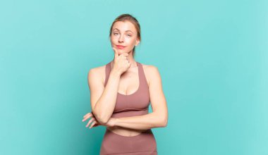 Genç sarışın kadın ciddi, düşünceli ve güvensiz görünüyor, bir kolu çapraz ve eli çenesinde, seçenekleri değerlendiriyor. spor konsepti