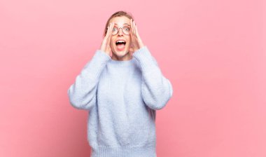 Genç, güzel sarışın kadın mutlu, heyecanlı ve pozitif hissediyor. Elleri ağızda, bağırıyor.