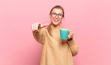 Genç sarışın kadın kameraya bakıyor, kendinden emin, arkadaş canlısı bir gülümsemeyle, seni seçiyor. kahve konsepti